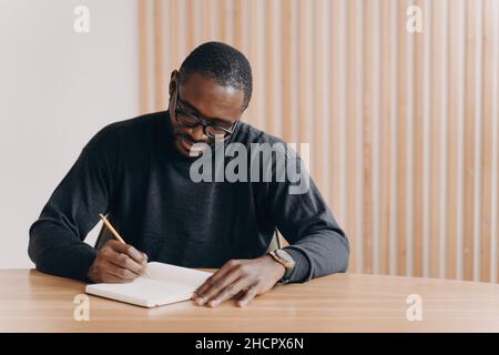 Fokussierter junger afrikanischer Mann in einer Brille, der den Stift hält, während er am Schreibtisch sitzt und sich Notizen zur Tagesordnung macht Stockfoto