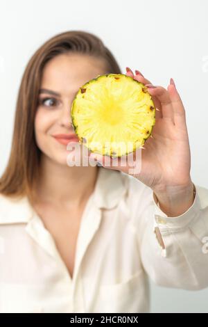 Eine junge, attraktive, hübsche, kaukasisch lächelnde Frau hält eine Ringschneide-Ananas, die ihr Auge vor einem weißen Hintergrund bedeckt. Gesunde Ernährung Konzept Stockfoto