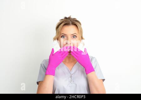 Das Porträt einer jungen kaukasischen Ärztin oder Krankenschwester ist schockiert, wenn sie ihren Mund mit ihren rosafarbenen Handschuhen vor weißem Hintergrund bedeckt Stockfoto