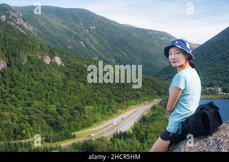 Eine chinesische Frau, die sich an einem großen Felsbrocken auf einem Künstlerbluff lehnt und an einem sonnigen Tag die Landschaft der Frakonia Notch in New hampshire betrachtet. Stockfoto