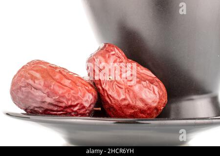 Zwei süße getrocknete ziziphus-Beeren auf einer Untertasse mit einer schwarzen Tasse, Nahaufnahme, isoliert auf Weiß. Stockfoto