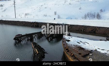 Luftaufnahme des Friedhofs von alten rostigen Schiffen in der Nähe der schneebedeckten Küste des kalten Meeres in der Wintersaison. Fliegen über zerstörte Boote und schneebedeckte Hügel in der Nähe von cal Stockfoto