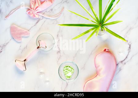 Gesichtsmassage. Feuchtigkeitscreme, rosa quarz Gesichtswalze und Gua Sha Stein auf Marmoroberfläche. Das grüne Pflanzenblatt ist zylindrisch. Stockfoto