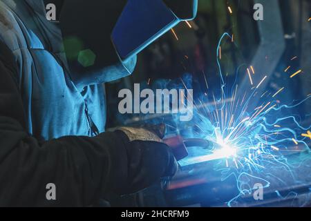 Starker Mann ist Schweißer, in einer Schweißmaske und schweißt Leder, ein Metallprodukt wird mit einem Schweißgerät in der Garage geschweißt, blaue Funken fliegen zum Stockfoto