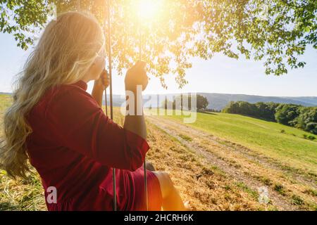 Rückansicht des glücklichen blonden Mädchens auf Schaukel im Sonnenlicht Stockfoto