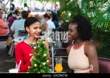 Zwei junge lateinische Frauen in eleganten Kleidern sitzen draußen und trinken ein erfrischendes Getränk Stockfoto