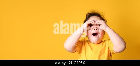 Kleine asiatische Mädchen macht Brille aus Fingern und lächelt, schaut auf die Kamera. Gelber Hintergrund. Leeres Leerzeichen Stockfoto