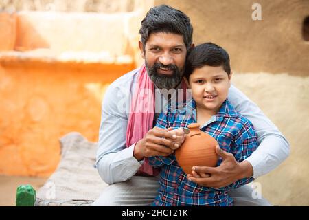 Vater und Sohn aus dem ländlichen Indien stecken Münzen in eine Tongeldkiste. Mann und Junge mit glücklichem Bart halten ein traditionelles Sparschwein oder Gulak, Vater lehrt das Kind zu sa Stockfoto