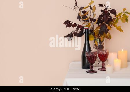 Vase mit Herbstzweigen, Flasche Wein, Gläsern und brennenden Kerzen auf dem Tisch in der Nähe der Lichtwand Stockfoto