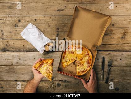 Männliche Hände halten ein Pizzateil mit Lieferkarton bereit, um es über einem Holztisch zu essen. Draufsicht Stockfoto