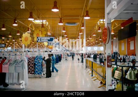 Das Innere des großen spanischen Supermarkts im Jahr 1977. Der riesige Lagerbereich umfasst Kleidung, die hier links zu sehen ist. Riesige, ausgeschnittene gelbe Blumen hängen von der Decke und die Farbpalette des Stores betont leuchtend gelb, rot und orange – die Grundfarben, die zu dieser Zeit beliebt waren. Dieses Bild stammt von einer Amateur-35mm-Farbtransparenz bei schlechten Lichtverhältnissen – einem Vintage-1970s-Foto. Stockfoto