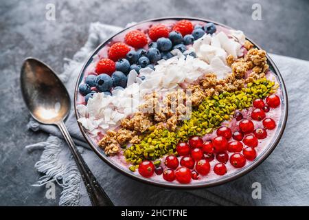 Gesunde Frühstückschale, mit Heidelbeer-Smoothie, Himbeere, Heidelbeere, Kokosnuss, Nüssen und Johannisbeeren auf einem Tisch mit Löffel Stockfoto