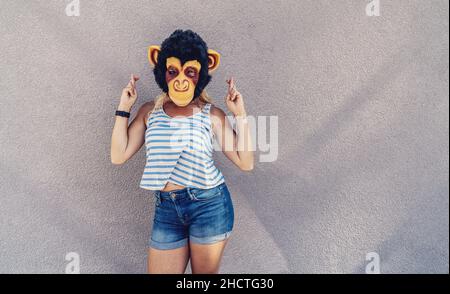 Verrückte junge Frau mit Affenmaske steht auf einer grauen Wand und Kreuzfinger. Copyspace für Ihren individuellen Text. Stockfoto