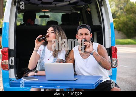 Das junge tätowierte Paar lächelt, trinkt Bier und benutzt den Laptop. Stockfoto