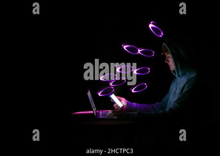 Ein junger Mann mit einer Kapuze, die auf sein Smartphone schaut. Menschen, die Technologie mit einem Laptop nutzen. Abstrakte rosa Lichtform im Hintergrund, zu Lichtmalerei gemacht. Stockfoto