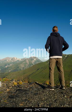 Ein Mann von hinten, mit Blick auf grüne Berge. Ein Wanderer betrachtet die Weiden und schneebedeckten Gipfel im Sommer in der Morgendämmerung. Stockfoto