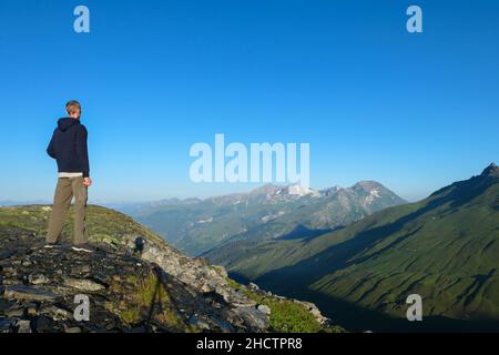 Ein Mann von hinten, mit Blick auf grüne Berge. Ein Wanderer betrachtet die Weiden und schneebedeckten Gipfel im Sommer in der Morgendämmerung. Stockfoto