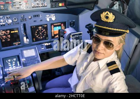 Schöne blonde Frau Pilot trägt Uniform und Hut mit goldenen Flügeln - moderne Flugzeug-Cockpit bereit zum Start Stockfoto