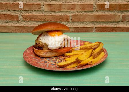 Großer Rindfleischburger mit geschmolzenem Käse, Spiegelei, Tomaten und Speck mit einer in Keile geschnittenen Seite von hausgemachten Pommes Stockfoto