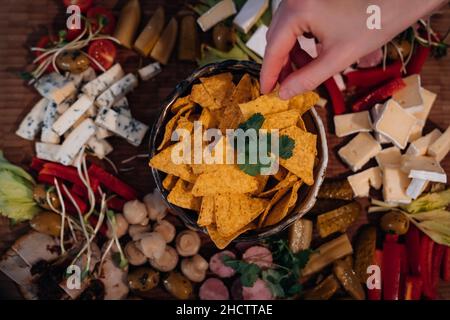Ein Bild einer Hand, die Nachos über dem Cold Cuts Board hält. Gurken, Oliven, Kirschtomaten, Sellerie-Sticks, Sprossen und andere gesunde Snacks. Stockfoto