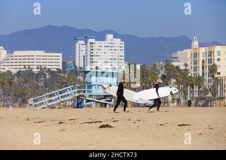 Surfer laufen den Strand vor einer Rettungsschwimmerhütte in Santa Monica, Kalifornien, USA, hinauf. Hotels in der Innenstadt im Hintergrund Stockfoto