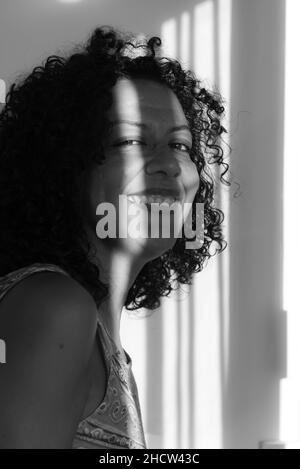 Schwarz-Weiß-Porträt des Gesichts einer Frau mit Licht und Schatten aus dem Fenster. Salvador, Bahia, Brasilien. Stockfoto