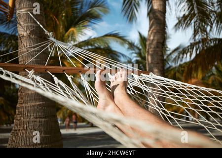 Beine eines jungen Mannes in der Hängematte. Touristen ruhen unter Kokospalmen am tropischen weißen Sandstrand. Stockfoto