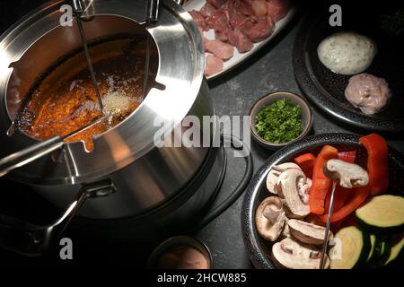 Fondue-Topf mit heißem Öl, langen Gabeln, Gemüse und rohem Fleisch, festliches Party-Dinner für Freunde und Familie, ausgewählter Fokus, enge Schärfentiefe Stockfoto