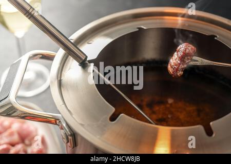 Knusprig gebratenes Stück Fleisch auf einer langen Gabel über einem Fondue-Topf mit heißem Öl, geselliges Abendessen, das oft zu Neujahr für Freunde und Familie serviert wird, ausgewählt f Stockfoto