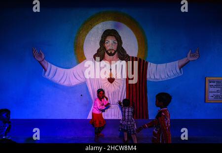 In Haulia, Westbengalen, Indien, feiern Kinder aus verschiedenen fernen Dörfern in einer kleinen Kirche das Weihnachtsfest und spielen vor einem großen Jesus Christus-Foto. Stockfoto