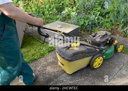 Körperteil des Mannes, der im Frühjahr den Rasenmäher durch einen kleinen Hinterhof schiebt. Niedriger Winkel Ansicht des Gärtners in grün insgesamt. Stockfoto