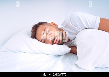Hübscher, netter Mann, der im Bett mit weißen Bettdecken schläft. Mann liegt auf dem Kissen und genießt einen guten gesunden Schlaf Stockfoto