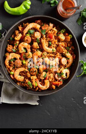 Kreolischer Jambalaya mit Huhn, geräucherten Würstchen und Gemüse in einer Pfanne auf schwarzem Stein. Draufsicht, flach liegend Stockfoto