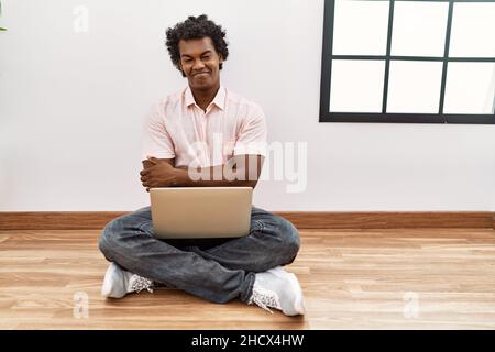 Afrikanischer Mann mit lockigen Haaren mit Laptop auf dem Boden sitzen glückliches Gesicht lächelnd mit gekreuzten Armen Blick auf die Kamera. Positive Person. Stockfoto