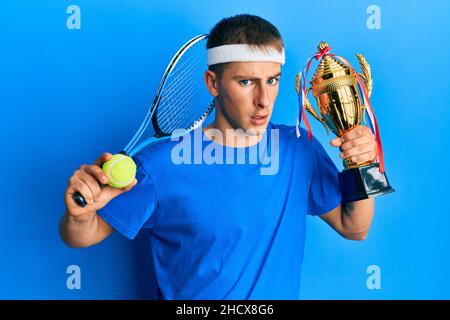 Junge kaukasische Mann spielt Tennis hält Trophäe ahnungslos und verwirrt Ausdruck. Zweifel Konzept. Stockfoto