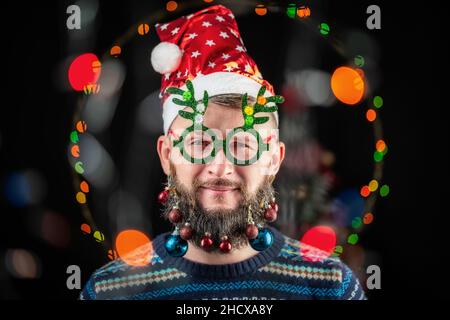 Ein Mann mit weihnachtsmannhut und dekorativem Spielzeug auf seinem Bart begrüßt in einem Kreis von farbigen Bokeh-Lichtern das gute Neue Jahr. Stockfoto