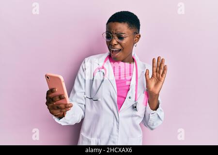 Junge afroamerikanische Ärztin, die Videoanruf mit dem Smartphone im Schockgesicht macht, skeptisch und sarkastisch aussieht, überrascht mit offenem Mund Stockfoto