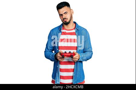 Junger hispanischer Mann mit Bart spielt Videospiel hält Controller ahnungslos und verwirrt Ausdruck. Zweifel Konzept. Stockfoto