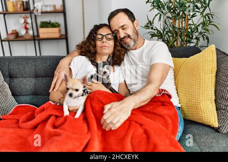 hispanische Paare mittleren Alters, die zu Hause mit Hunden auf dem Sofa sitzen. Stockfoto