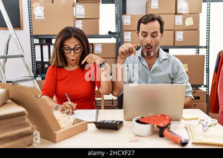 Ehepaar im mittleren Alter, das bei einer E-Commerce-Firma in kleinen Unternehmen arbeitete, zeigte nach unten mit Fingern, die Werbung zeigten, überraschte das Gesicht und den offenen Mund Stockfoto