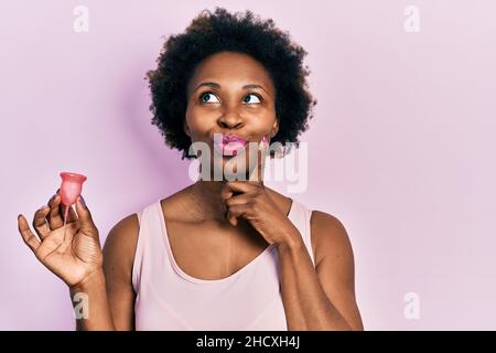 Junge afroamerikanische Frau hält Menstruationstasse ernstes Gesicht Denken über Frage mit Hand auf Kinn, nachdenklich über verwirrende Idee Stockfoto