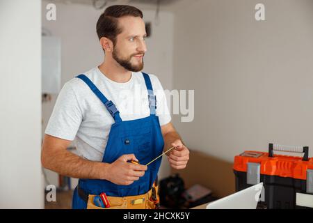 Fröhlicher Reparaturmitarbeiter in Uniform, der Werkzeuggürtel trägt, Maßband hält und im Gebäude wegschaut Stockfoto
