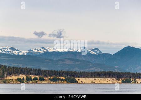 Ein Blick von der Straße von Georgia auf die ökologisch sensiblen Komas Bluffs von Denman Island mit dem Comox Glacier von Vancouver Island im Hintergrund. Stockfoto