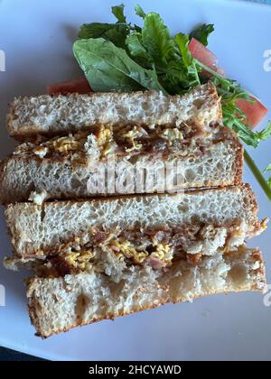 Zwei Thunfisch-Ei-Sandwiches auf einem Teller mit Tomaten und grünem Salat. Die Füllung des Sandwiches besteht aus Thunfisch-Stücken und hart gekochten Eiern aus der Nähe Stockfoto