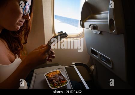 Kaukasische Frau, die in der Economy Class fliegt und an Bord des Flugzeugs ein Mittagessen von einem Tablett isst. Stockfoto