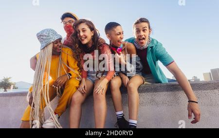 Multiethnische Gruppe von Freunden, die eine gute Zeit im Freien genießen. Fröhliche Freunde lachen fröhlich, während sie an einer Wand in der Stadt sitzen. Jugendliche mit f Stockfoto