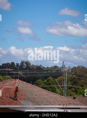 Mann, der im Surburban Sydney mit einem Hochdruckreiniger Flechten und Moos vom Terrakotta-Ziegeldach reinigte Stockfoto