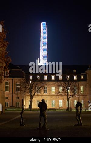 Stuttgart, 31. Dezember 2021: Riesenrad hinter dem Schlossplatz. Die Gebäudefassade ist nachts orange beleuchtet. 3 Skulpturen im Vordergrund. Stockfoto