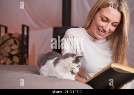 Eine erfreuliche Frau las ein Buch mit einer Katze an ihrer Seite Stockfoto