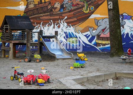 Kinderspielplatz in einer Stadt, mit zahlreichen Spielsachen, Sandkasten und Rutsche vor einer Wand mit bunten Straßenkunst Stockfoto